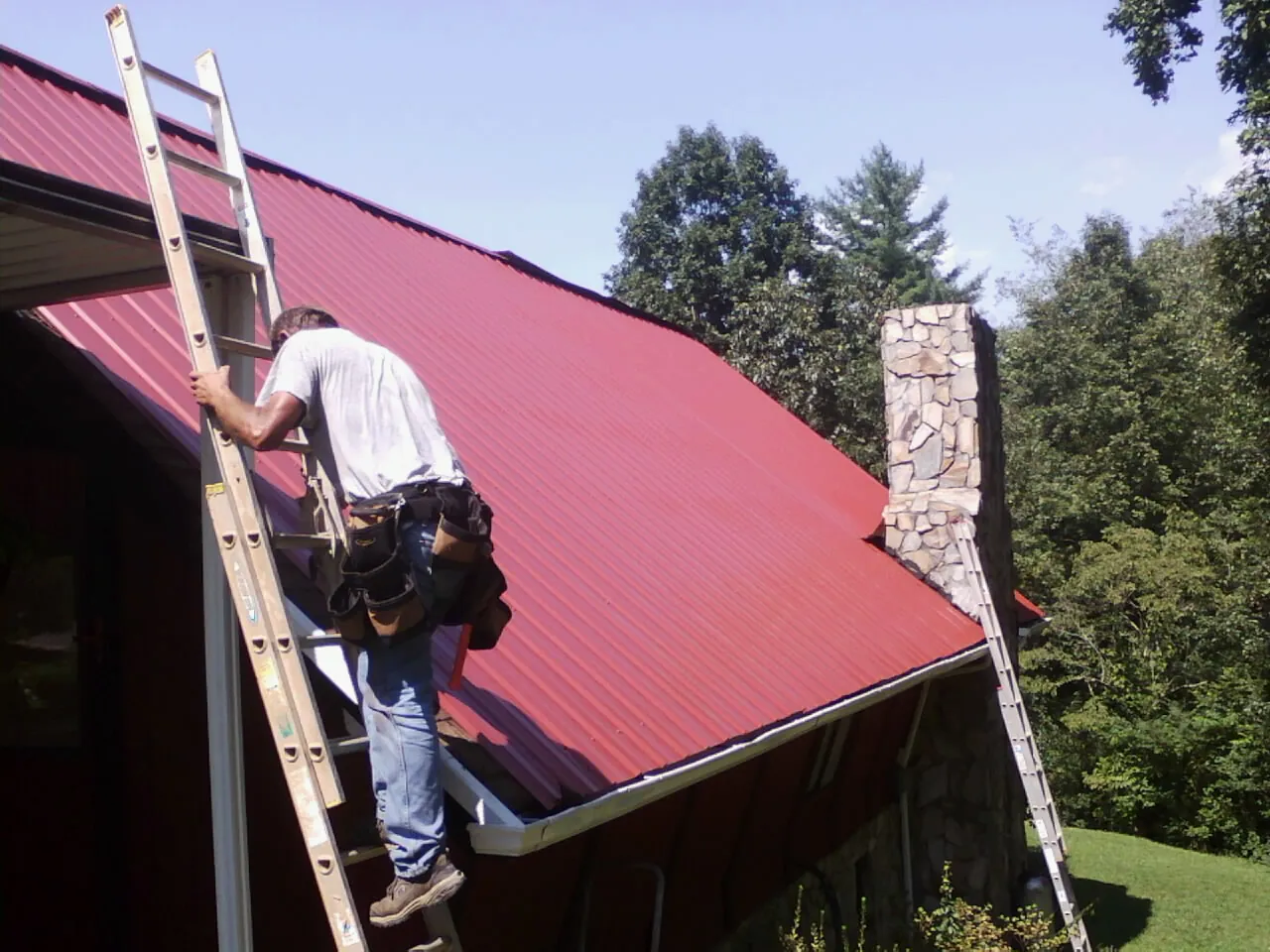 Roofer on a ladder beside a red metal roof and stone chimney — Soffit & Fascia Repair by Roofing Brothers in Laceyville